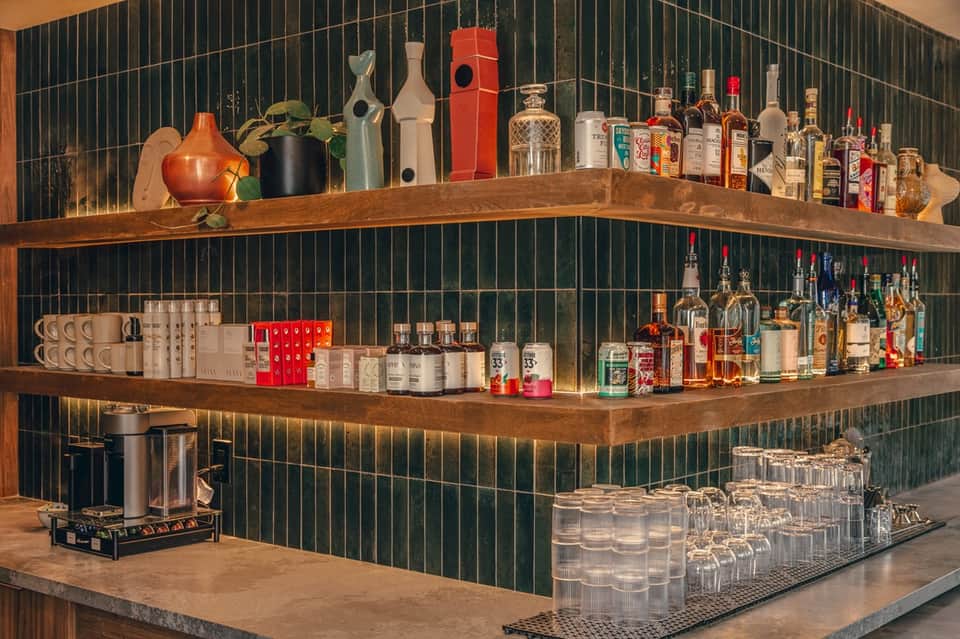 Modern bar corner with curved wooden shelves, green tile backsplash, spirits display, and decorative items