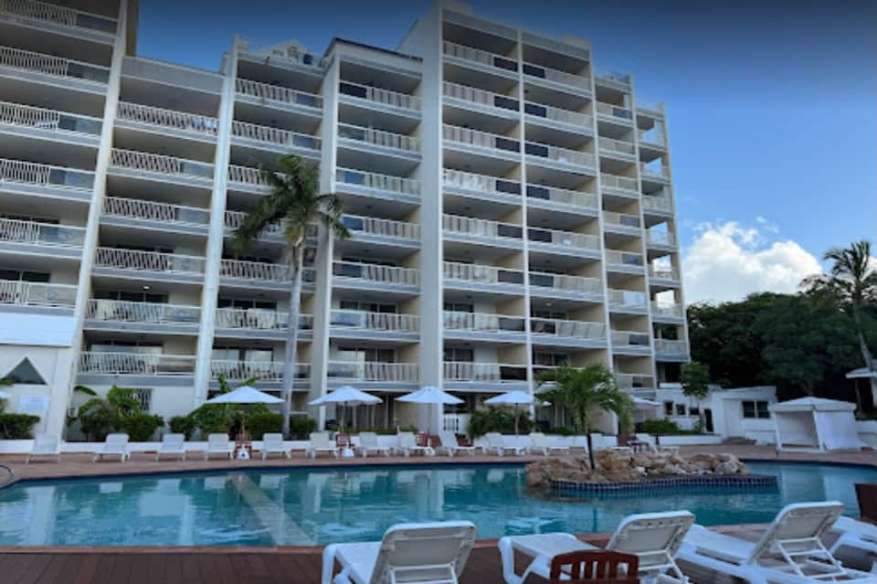 Oceanfront hotel building with balconies overlooking pool deck and blue umbrellas by the water