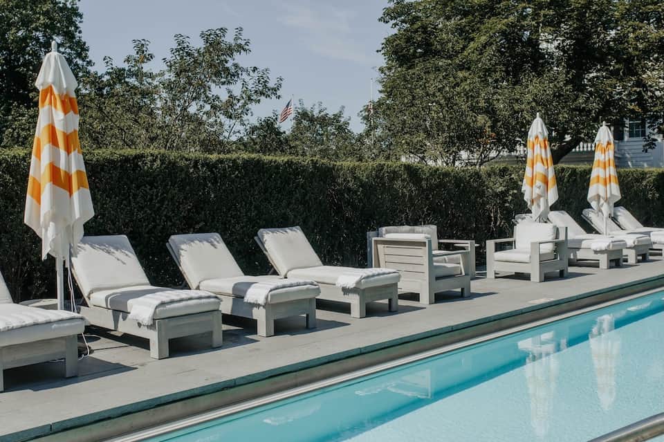 Row of white lounge chairs with striped umbrellas along turquoise pool, with hedge wall and clear sky