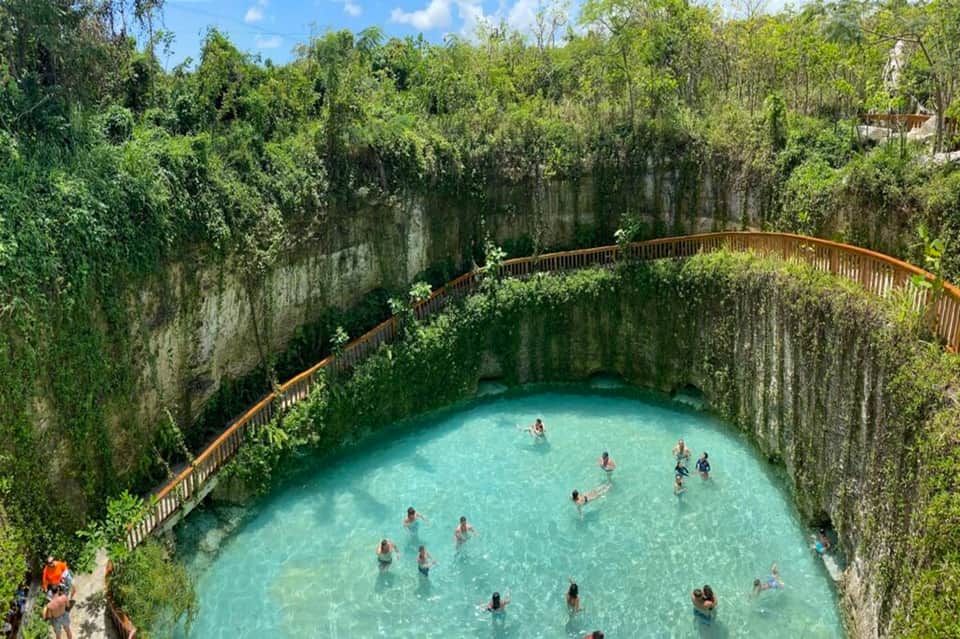 Cenote natural sinkhole pool surrounded by limestone cliffs, lush vegetation, and wooden railings with swimmers