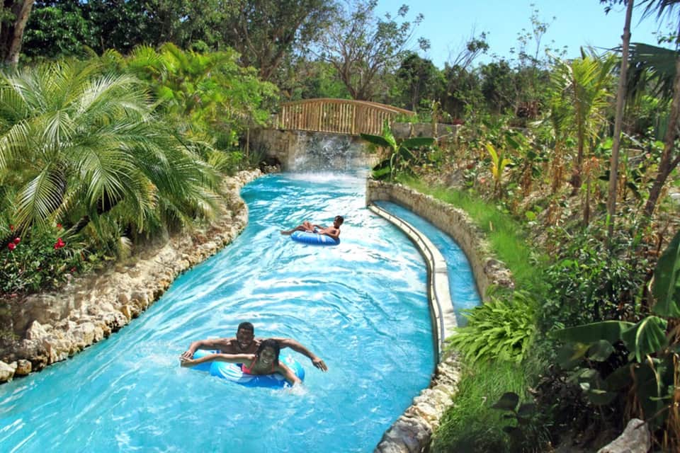 Turquoise lazy river with guests floating on tubes, surrounded by tropical vegetation and stone walls