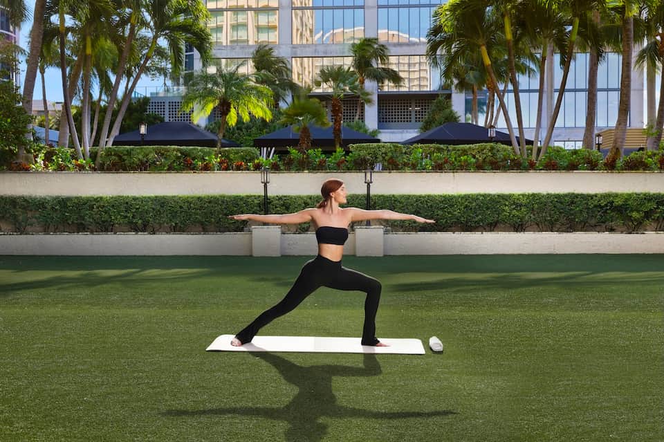 Woman performing yoga pose on grass patio with palm trees and downtown skyline backdrop