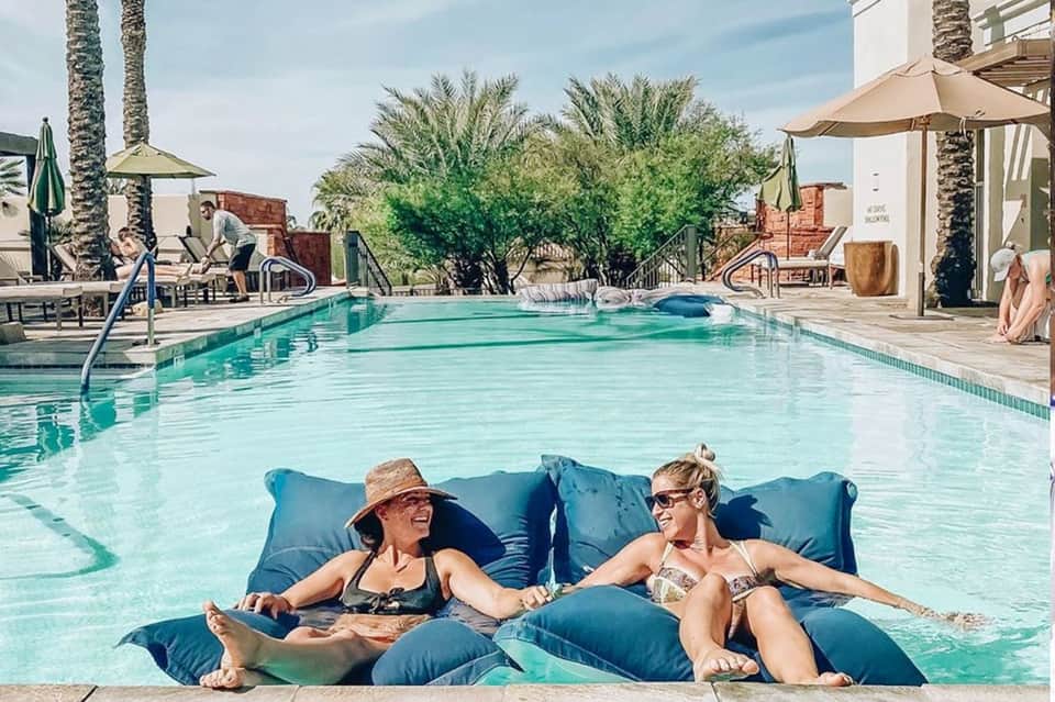 Two guests relaxing on blue loungers in a resort pool surrounded by palm trees and umbrellas