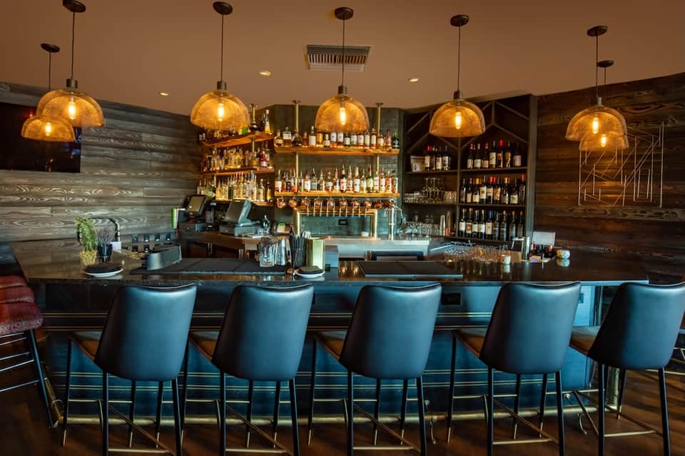 Upscale bar with golden dome pendant lights, dark wood shelving, blue bar stools, and glass backlit counter