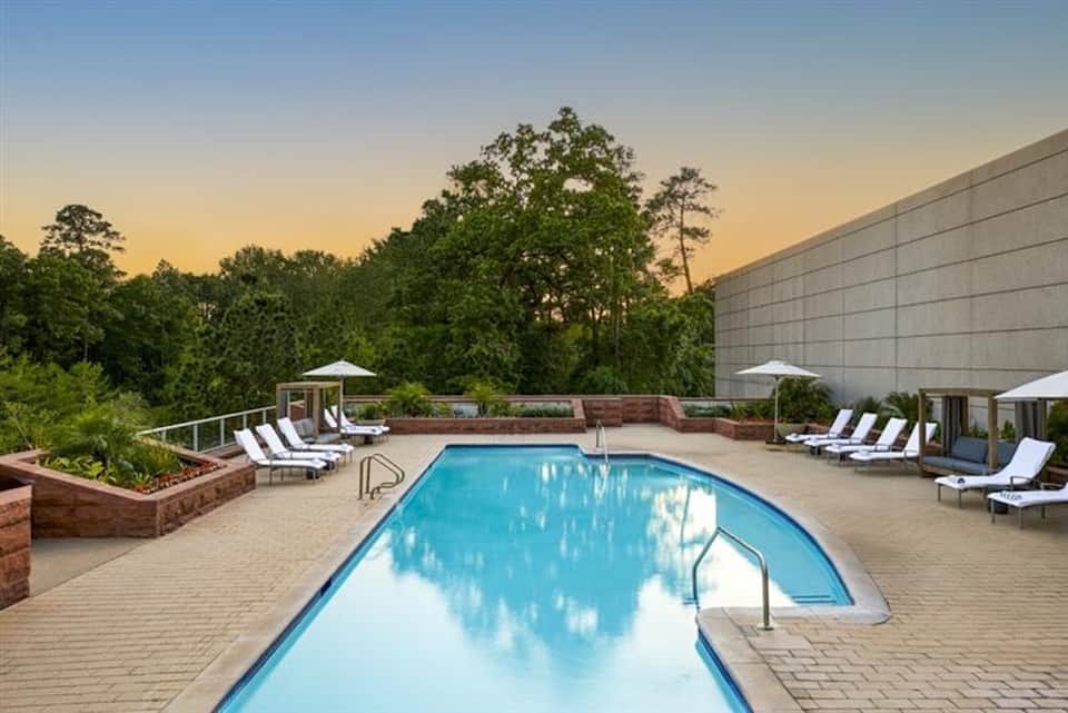 Hotel pool at dusk with lounge chairs, white umbrellas, and forest backdrop at sunset