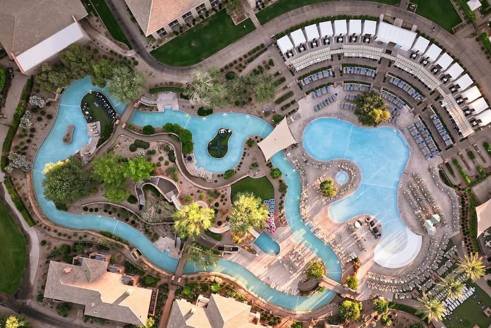 Aerial view of large resort complex with multiple curved pools, lounger arrangements, green landscaping, and parking area