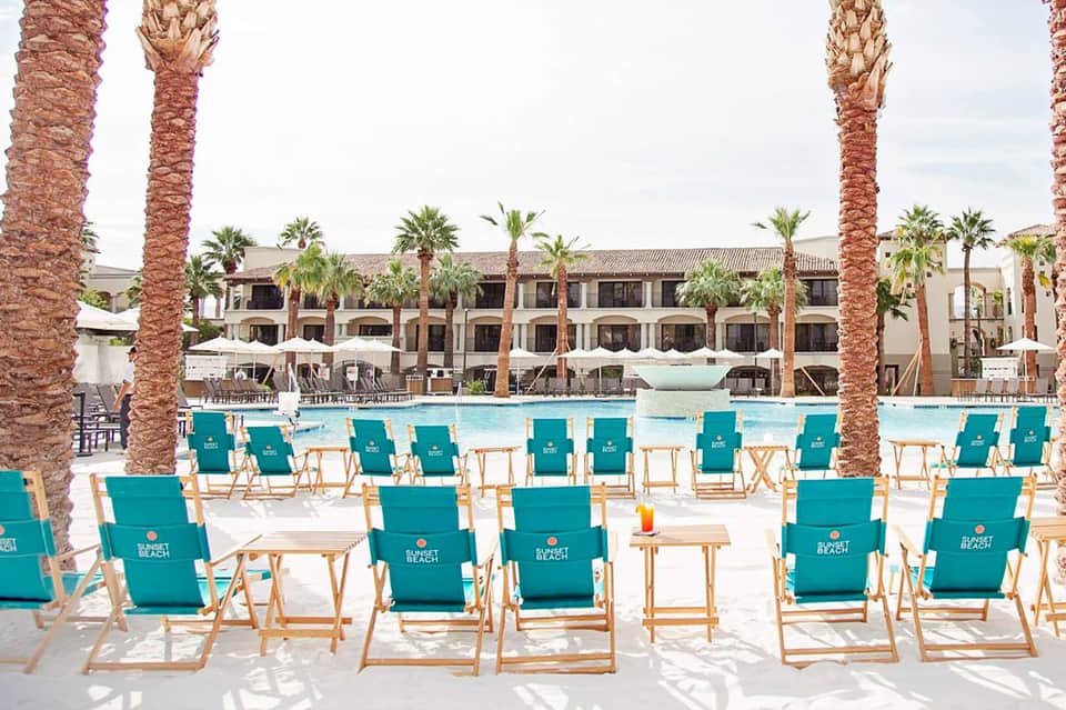 Resort pool area with turquoise lounge chairs, palm trees, and beachfront hotel building