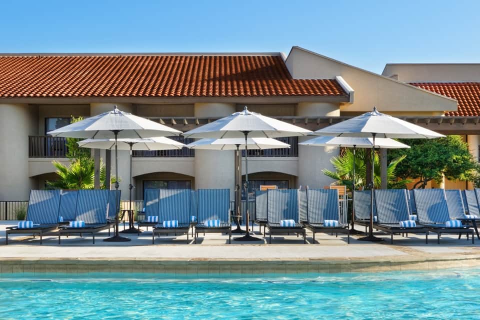 Resort poolside with white umbrellas, blue lounge chairs, clay tile roof building, and palm trees