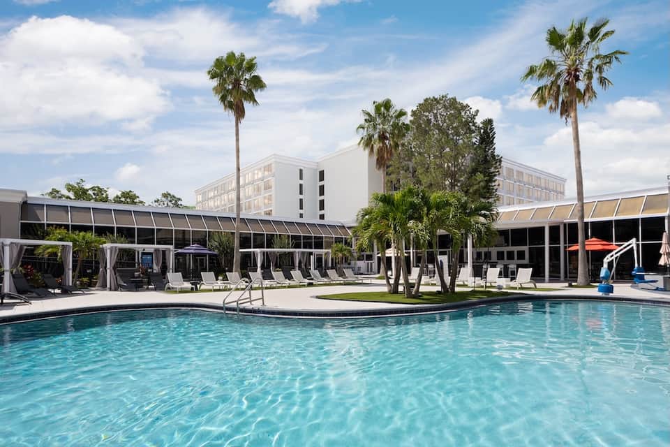 Resort pool with lounge chairs and umbrellas, palm trees, white modern hotel building, and covered cabanas
