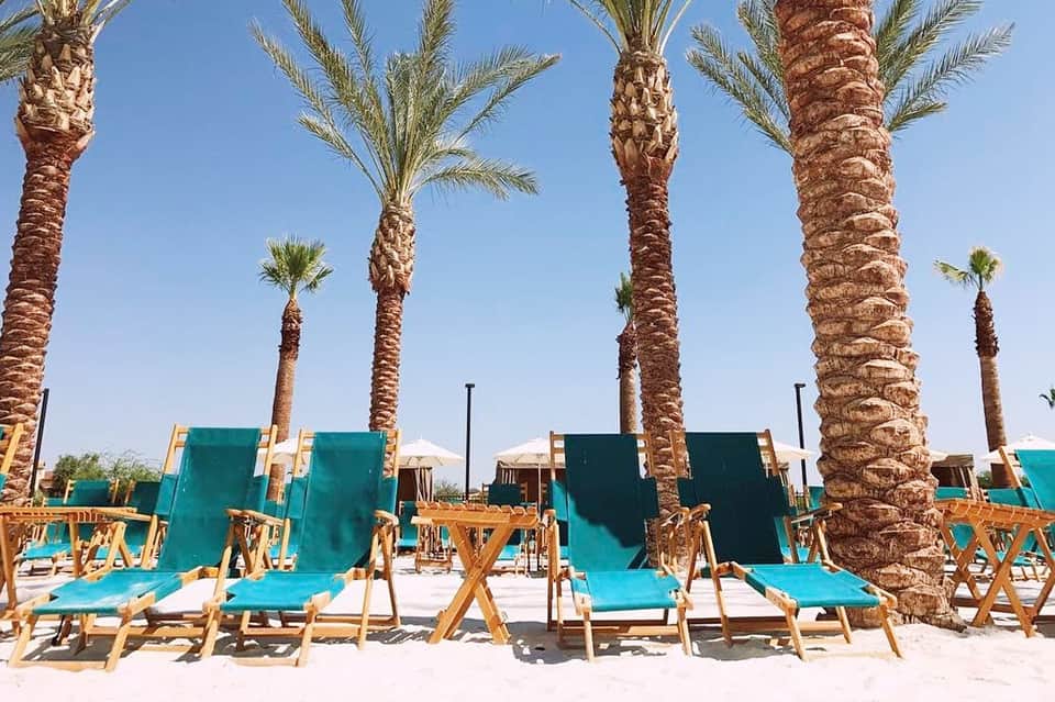 Beach lounge area with turquoise deck chairs and tall palm trees against clear blue sky