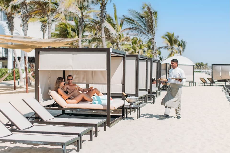 Beachfront cabana beds with sun shades on sandy beach lined with palm trees and white umbrellas
