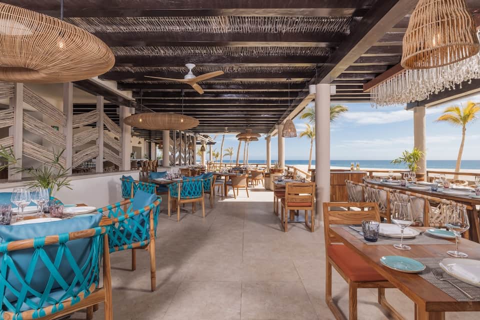 Upscale beachfront restaurant with ocean views, woven pendant lights, turquoise accent chairs, and modern tropical design