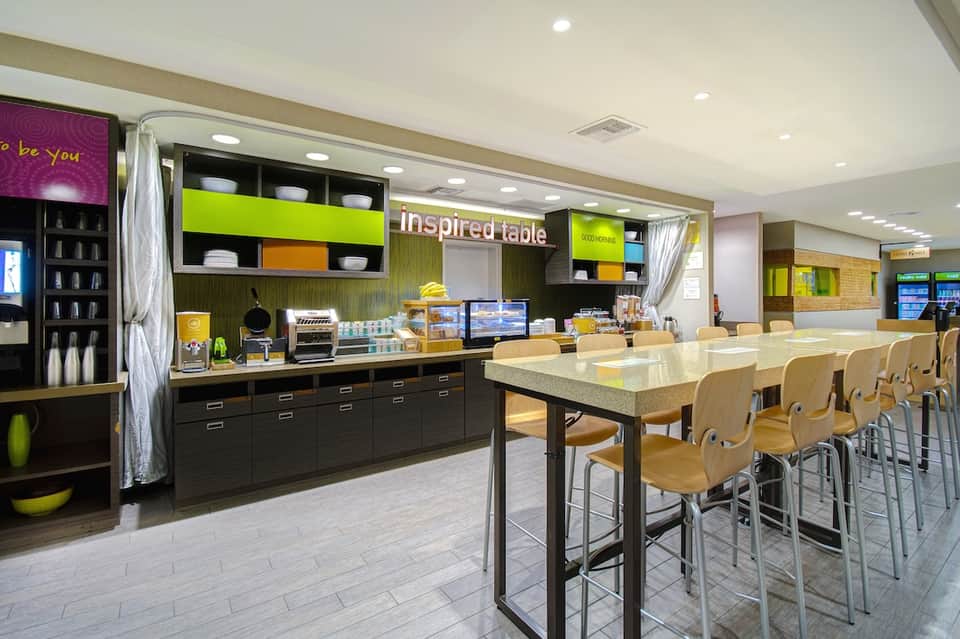 Hotel breakfast room with green accents, counter service, and high-top seating areas