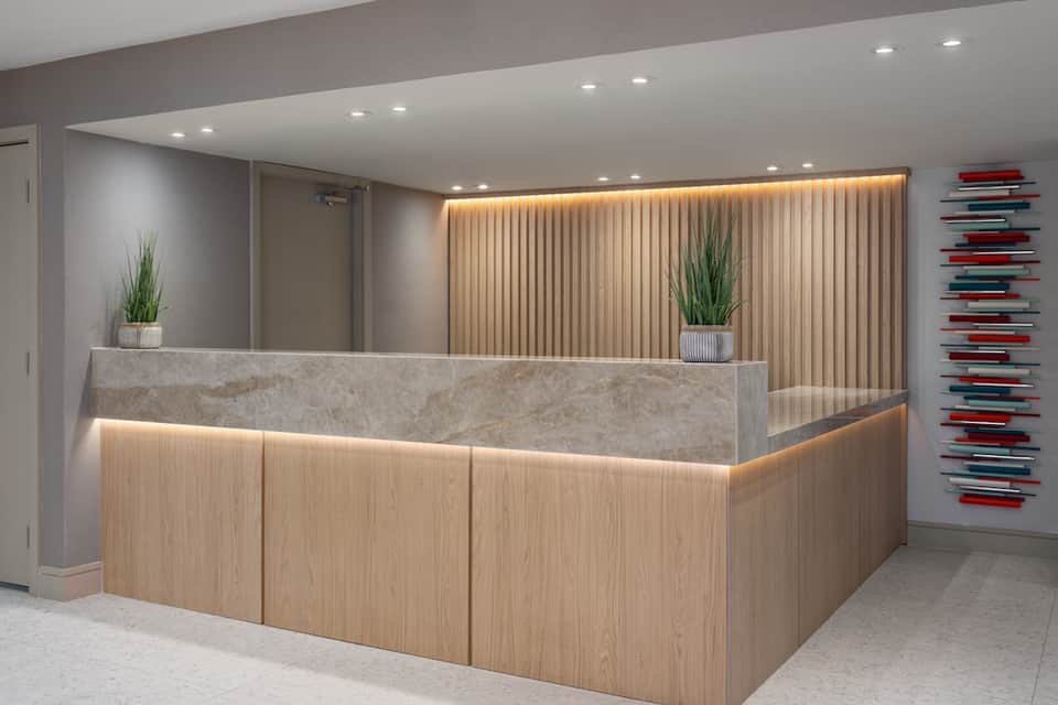 Modern hotel reception desk with warm wood paneling, stone countertop, ambient lighting, and potted plants
