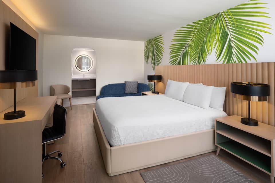 Contemporary hotel room with queen bed, palm leaf accent wall, and workspace area