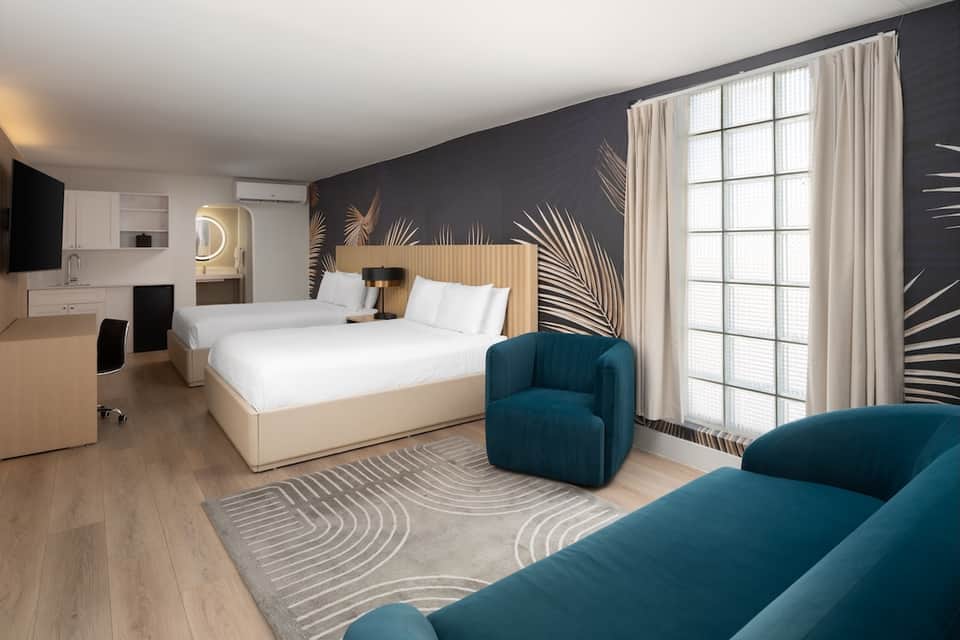 Modern hotel bedroom with botanical wall mural, teal accent chair, warm wood furnishings, and floor-to-ceiling windows