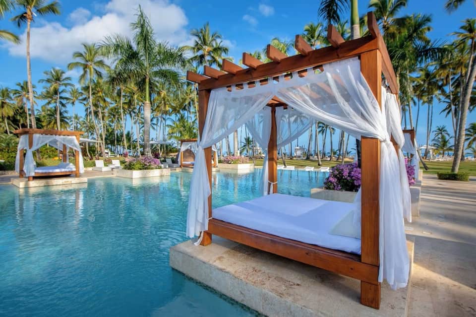 Elegant resort pool with wooden cabana, white curtains, palm trees, and ocean view in tropical setting