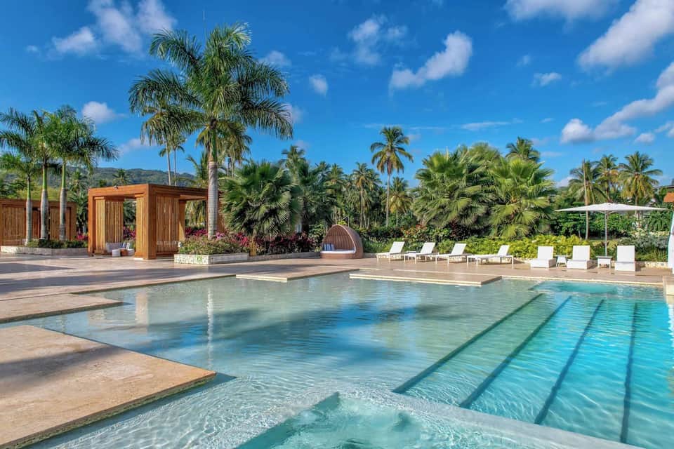 Resort swimming pool surrounded by palm trees, wooden pavilion, white loungers, and tropical landscaping under blue sky