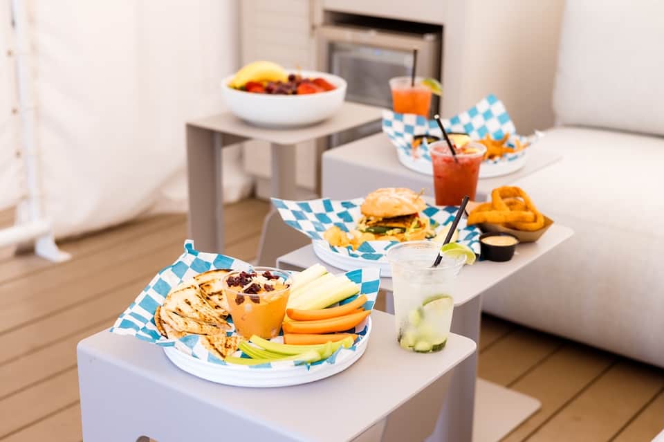 Outdoor dining setup with white plates, blue patterned napkins, burgers, beverages, and fresh fruit