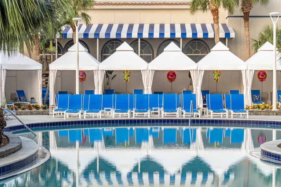 Resort pool deck with white cabanas, blue lounge chairs, palm trees, and striped awning