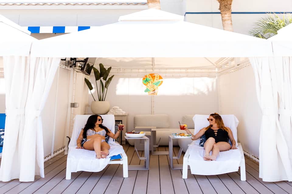 Two women lounging in white cabana beds under flowing white curtains and palm umbrella