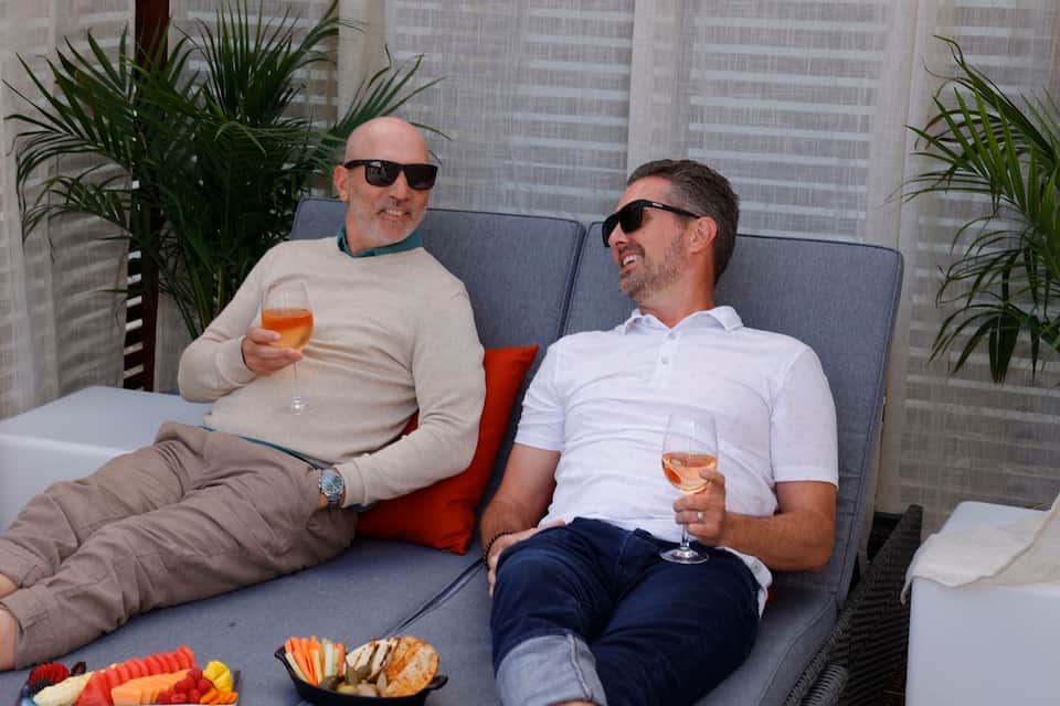 Two men in sunglasses relaxing on lounge chairs with drinks and fruit platters on rooftop