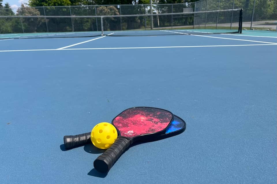 Pickleball paddle and yellow ball on blue court with net and fencing in outdoor recreation area