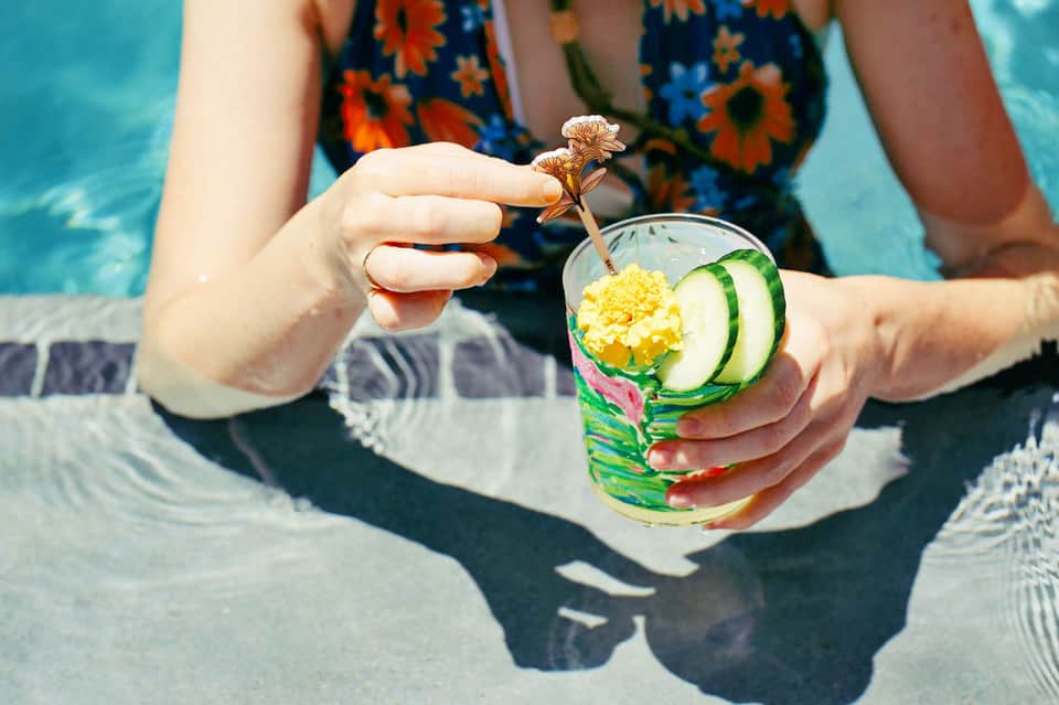 Person holding tropical cocktail with cucumber and flower garnish by pool