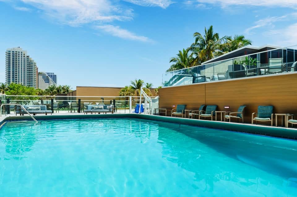 Modern resort pool with turquoise water, wooden deck, palm trees, and city skyline backdrop