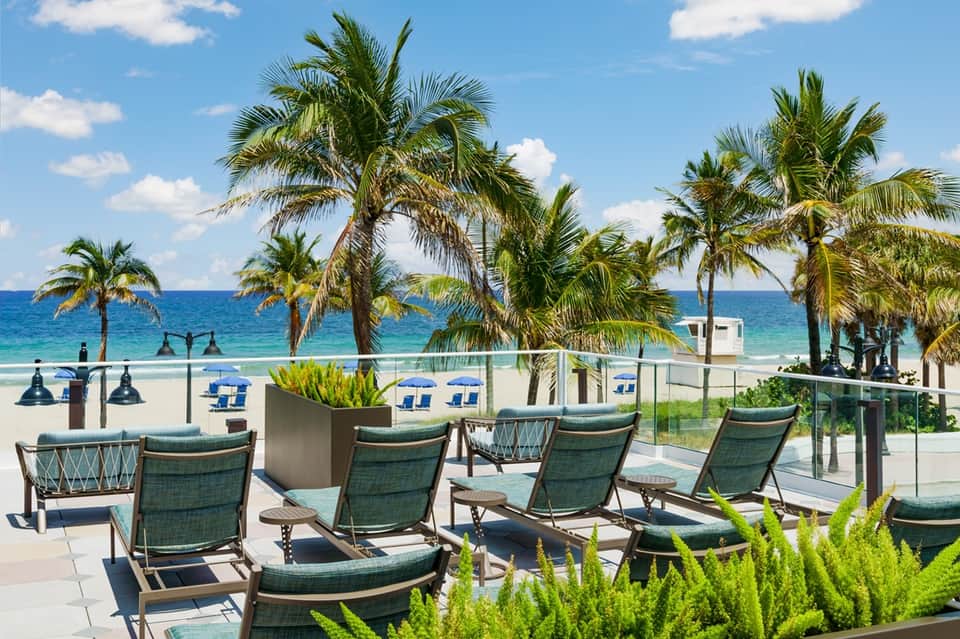 Beachfront deck with lounge chairs, palm trees, ocean view, and blue umbrellas on the beach