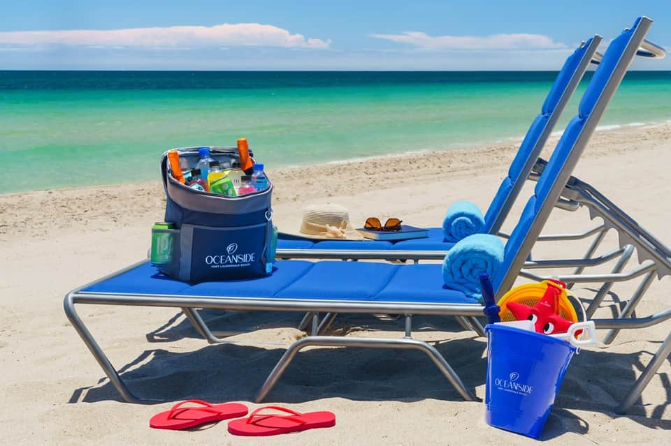 Beach lounge chairs with blue cushions, branded coolers, and colorful beach toys on sandy shore with ocean