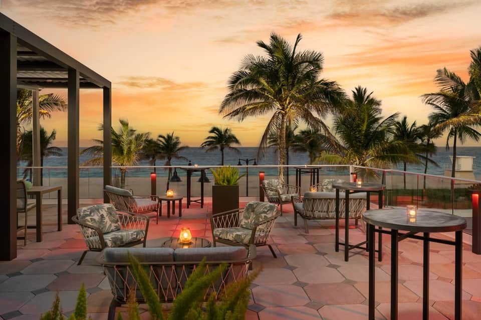 Beachfront resort patio at sunset with lounge chairs, fire pit, palm trees, and ocean views