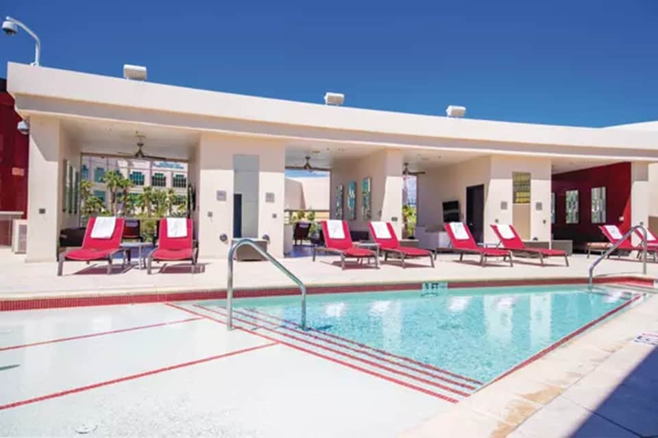 Hotel pool with pink and white loungers on covered patio near facility building
