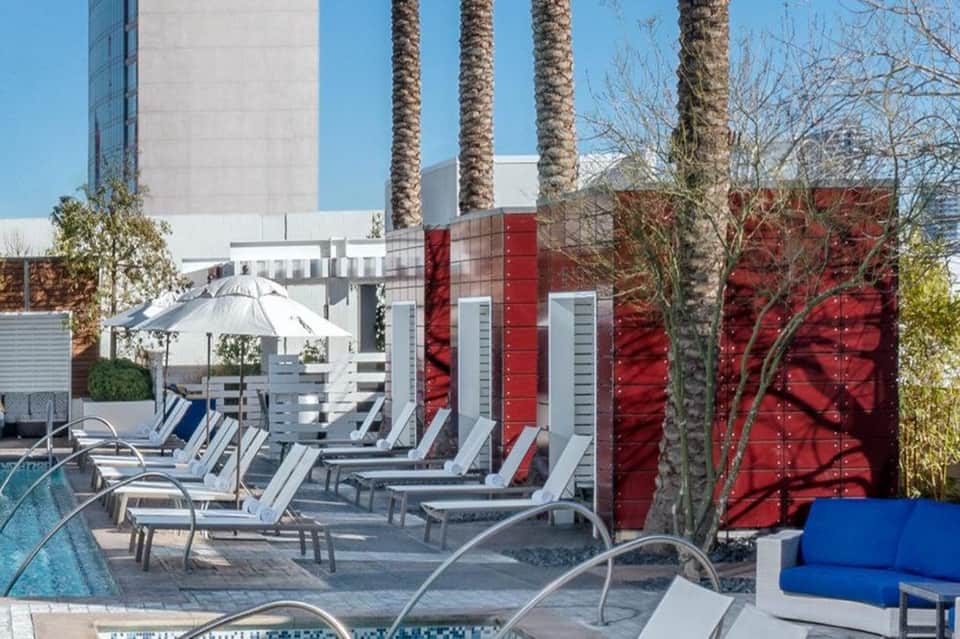 Resort pool deck with white lounge chairs, umbrellas, palm trees, and red privacy screens