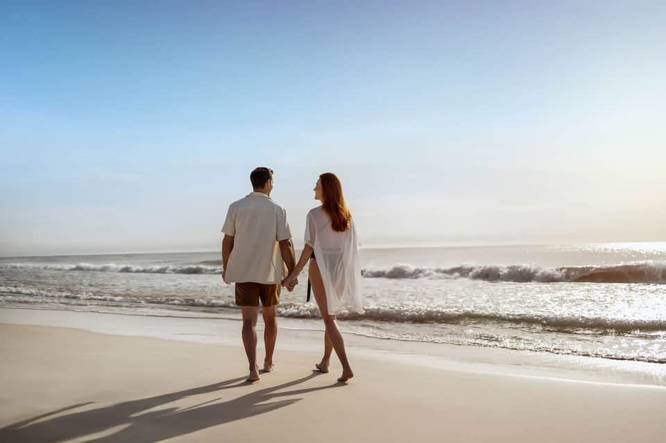 Couple holding hands walking on sandy beach at sunset with waves and clear sky in the background