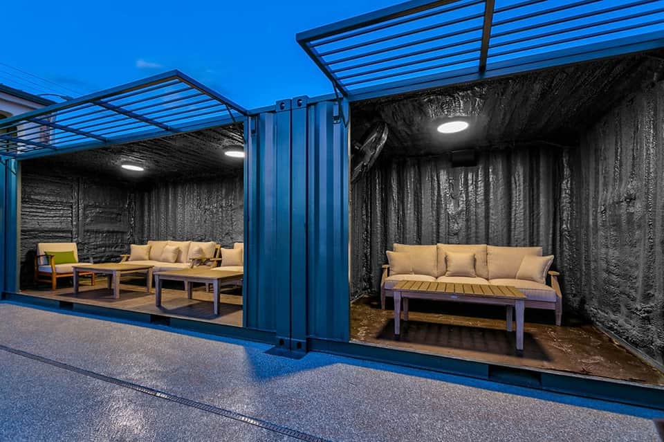 Modern outdoor lounge area with blue metal structure, black curtains, and beige cushioned seating on paved flooring.