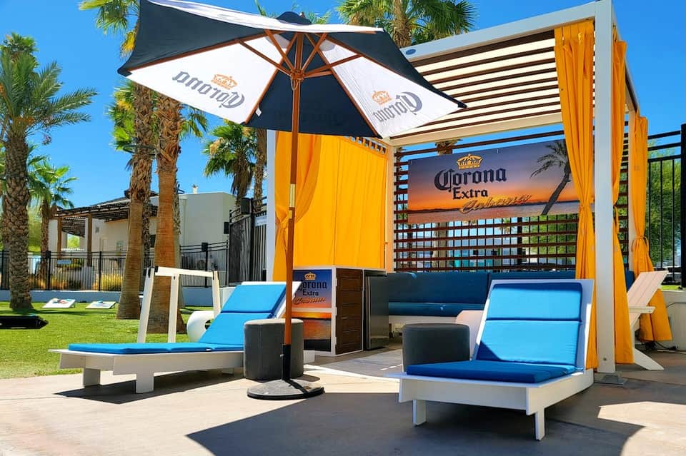 Poolside cabana with Corona branding, blue lounge chairs, yellow curtains, and palm trees