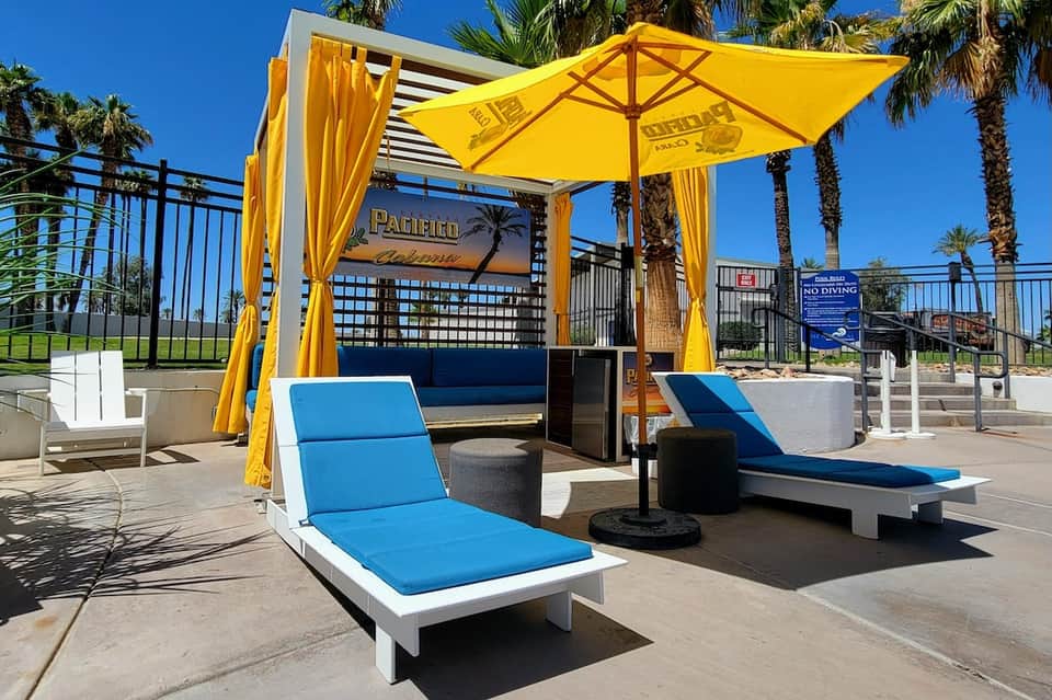 Poolside lounge area with yellow umbrella, blue cushioned seating, palm trees, and water view