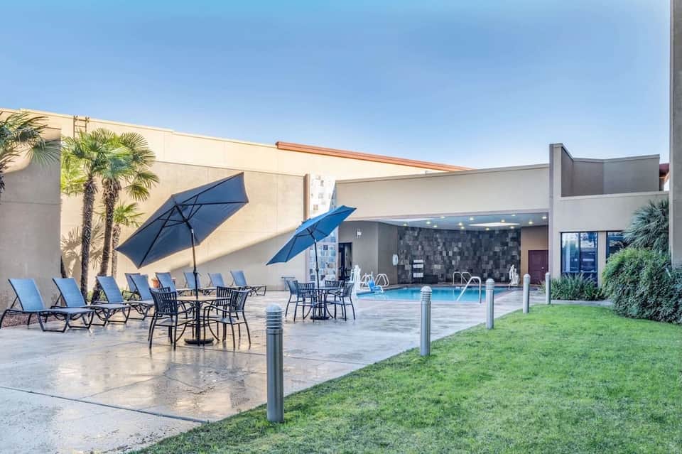 Modern resort pool area with blue umbrellas, lounge chairs, white walls, and landscaped grounds
