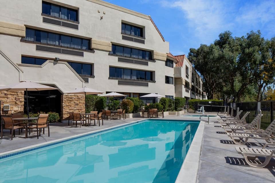 Resort pool with loungers, umbrellas, building facade, trees, and stone patio during daytime