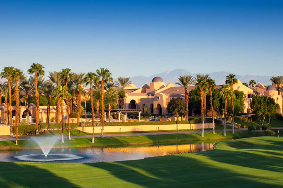 Luxury golf resort with palm trees, manicured green lawn, fountain, and cream-colored buildings with mountains in background