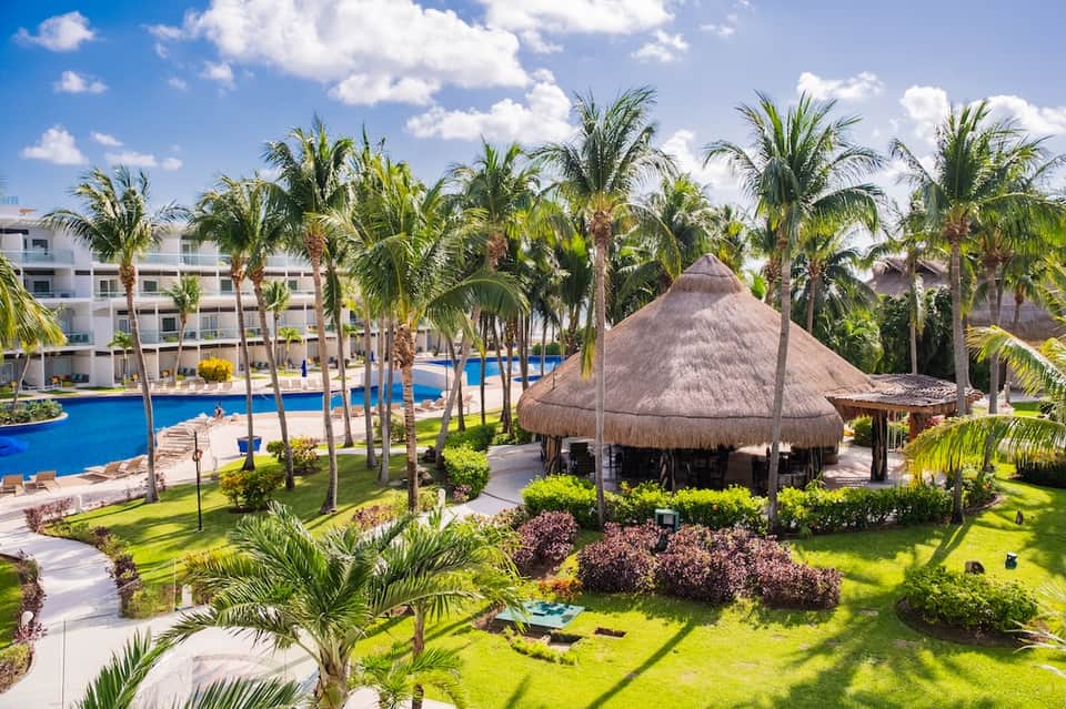 Tropical resort pool area with palm trees, thatched-roof palapa, and beachfront accommodations