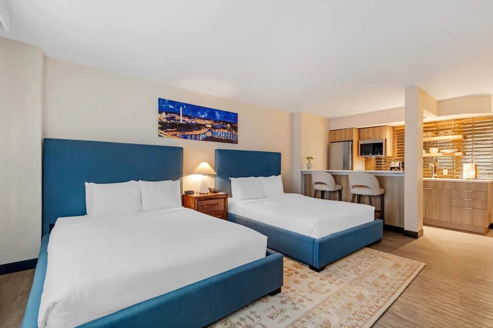 Hotel room with two queen beds with blue headboards, kitchenette, and contemporary décor