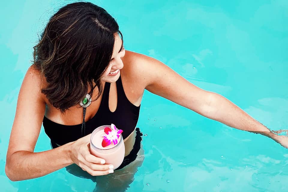 Woman in black swimsuit holding a tropical cocktail in turquoise pool water