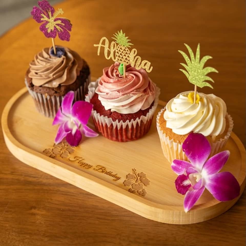 Three decorated cupcakes with tropical theme featuring orchids, pineapple topper, and birthday message on wooden board