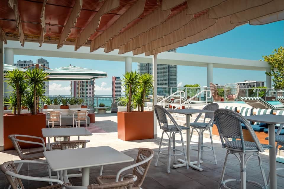 Modern rooftop terrace with dining tables, shade structures, ocean views, and high-rise buildings