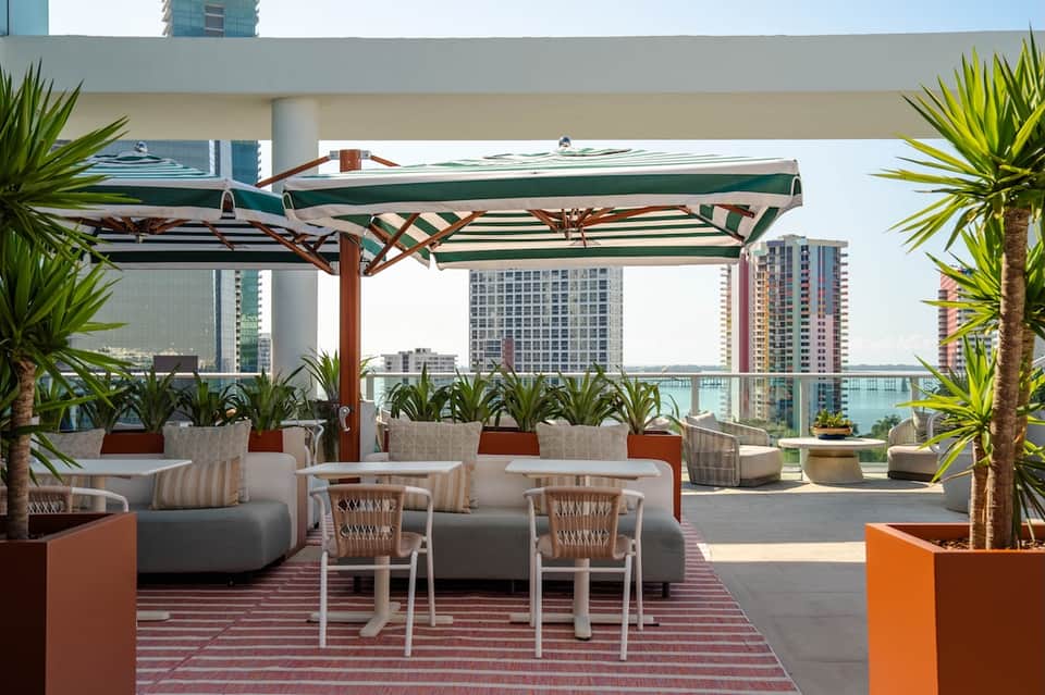 Rooftop lounge area with green striped umbrellas, seating, tropical plants, and city/waterfront views