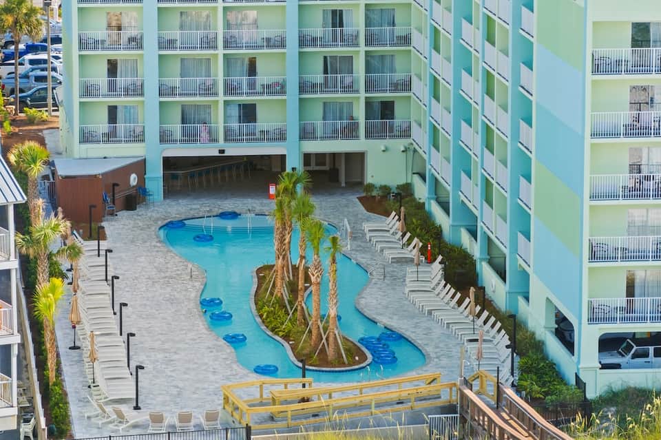 Aerial view of resort pool complex with blue water features, palm trees, and pastel-colored multi-story buildings