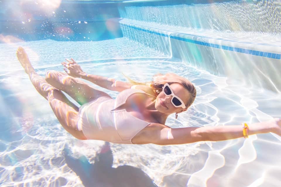 Woman floating underwater in swimming pool wearing white swimsuit and sunglasses