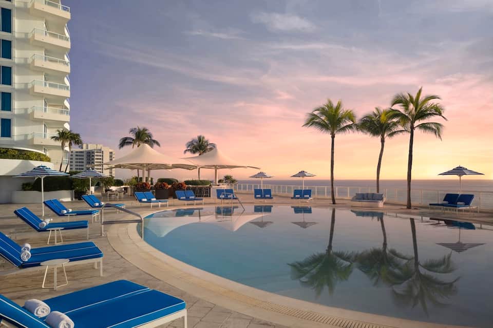 Beachfront pool at sunset with palm trees, blue loungers, umbrellas, and high-rise hotel